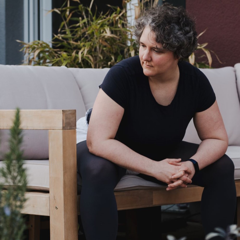 Katharina sitzt breitbeinig auf einer Bank. Sie hat die Ellenbogen auf die Knie gestützt und schaut diagnoal zu Boden. 